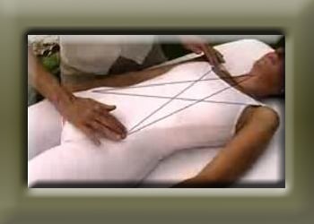 Woman in white body suit laying face up while man places tap across her for polarity therapy.