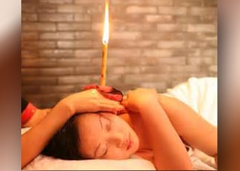 Woman holds a lit ear candling cone in the ear of a woman lying on her side