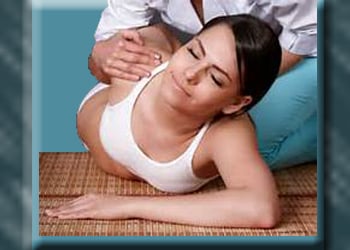 Woman in yoga clothing is laying on mat receiving swe thai massage from therapist crouching behind and pulling up on her right shoulder.