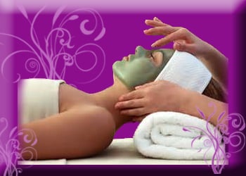 Woman laying face up with green mask on her face, white towel around her hair and woman's hands moving over her face.